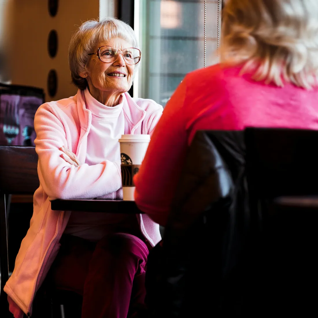 A senior woman in glasses chats warmly with a companion at a window-side cafe table