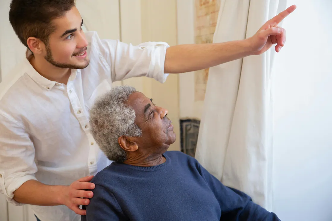 A young caregiver gently points something out to an elderly man, sharing the moment with patience