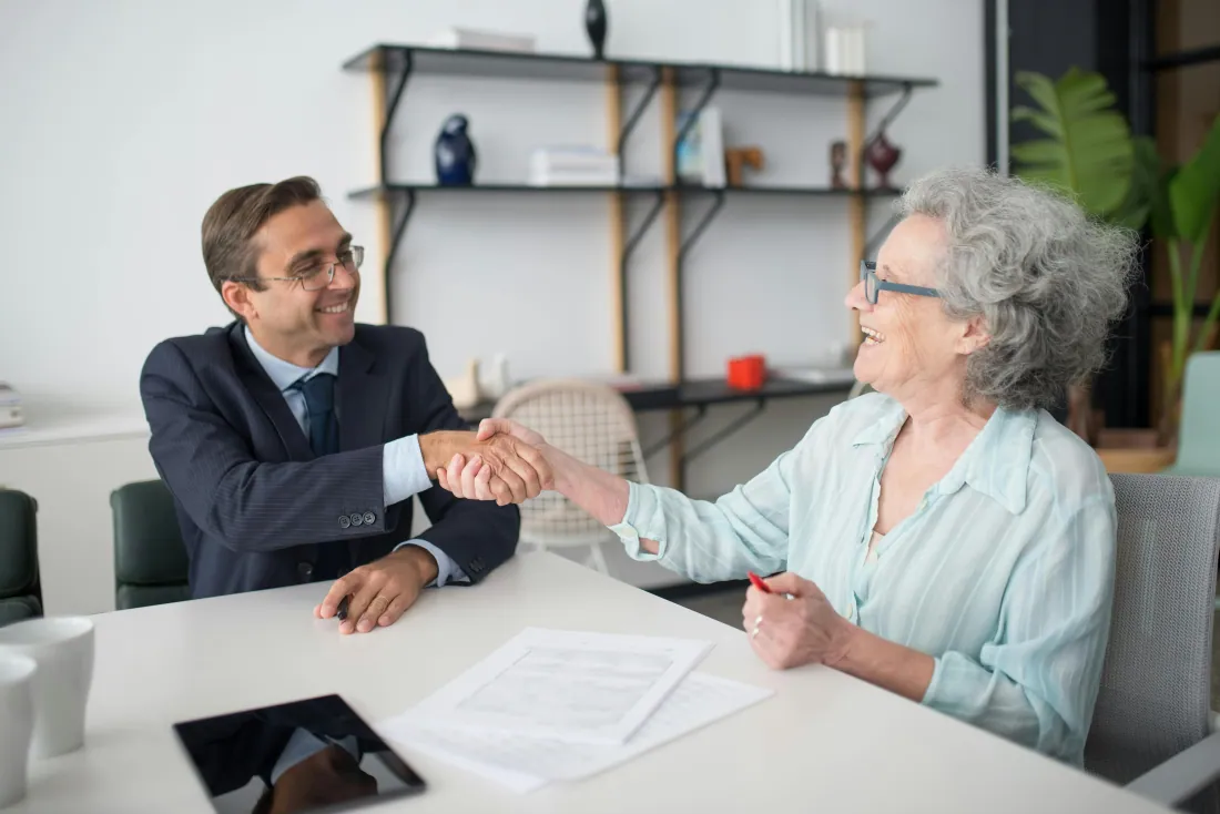 A senior woman and a trusted advisor share a warm handshake across a desk